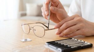 日本人の指先が、精密ドライバーを使い眼鏡のヒンジにある極小のネジを慎重に回す様子。机の上には精密ドライバーセットと小さな電池がある。