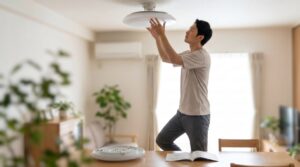 日本人の男性が、自宅のリビングルームで、脚立を使い安全に天井のLEDシーリングライトを交換する様子。資格不要な「軽微な工事」の範囲で、取扱説明書に従って作業している。