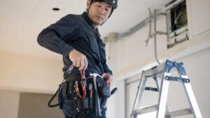 腰袋と安全コード付きの工具を装着した日本人作業者が、脚立そばで高所作業の準備をしている様子